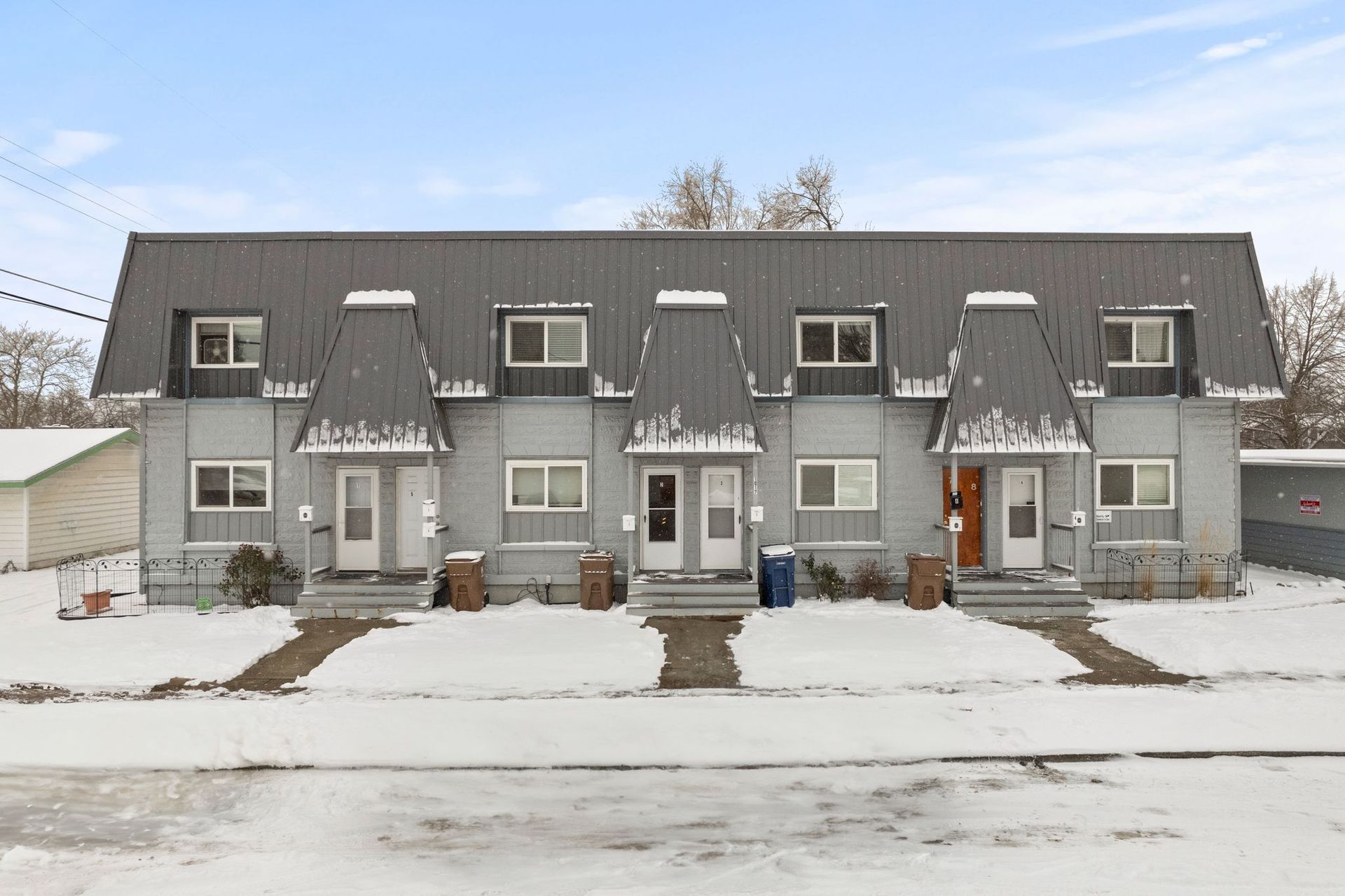 Row of gray townhouses with snow-covered roofs and walkways, set in a snowy landscape.
