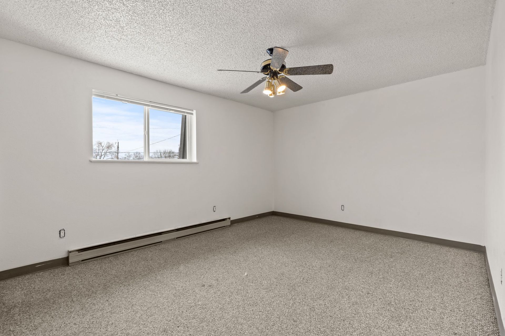 Empty room with a window, ceiling fan, and beige carpet. White walls, heater, and textured ceiling.
