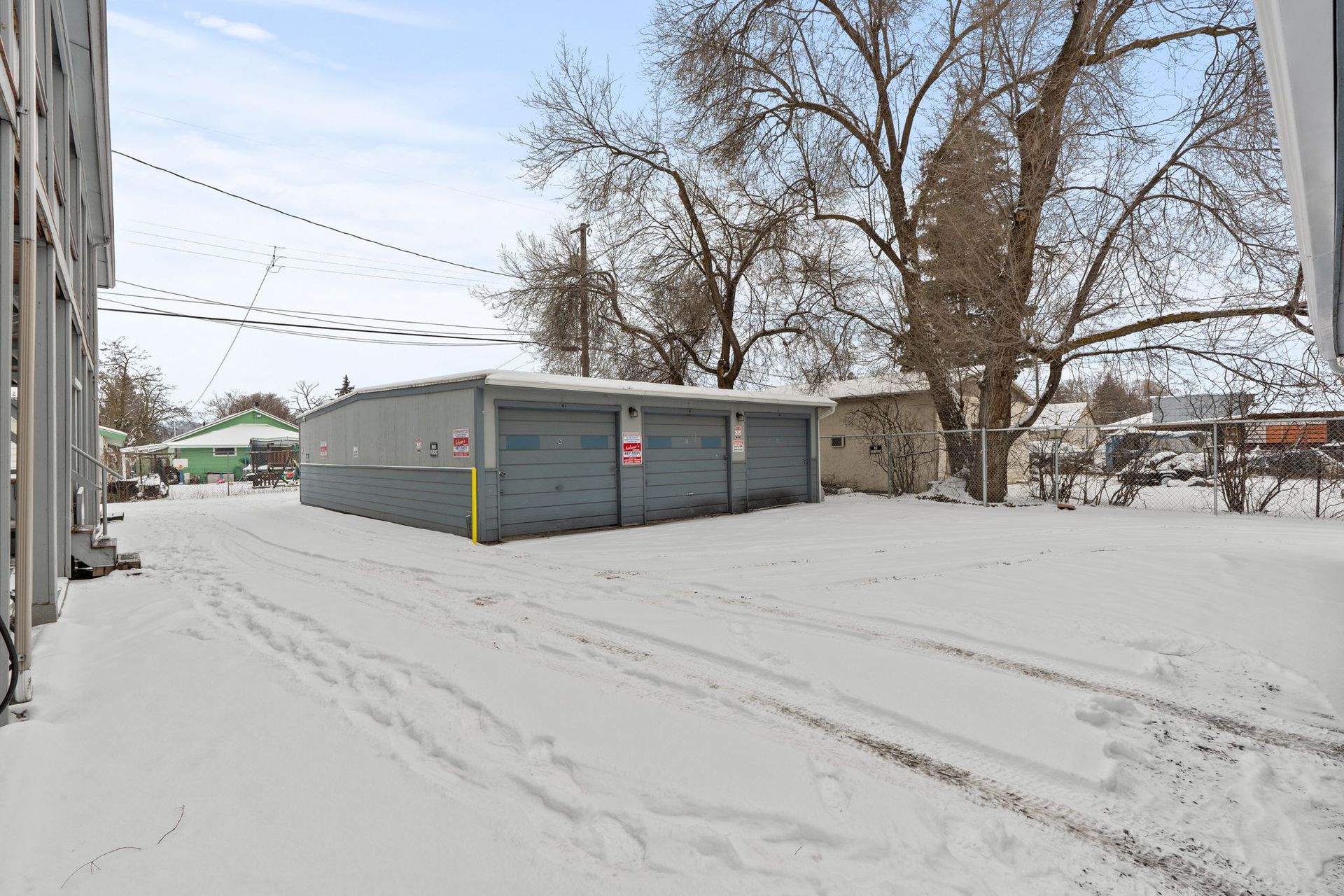 Snowy alley with three gray garages. Overhead power lines, bare trees, and a green building are visible.