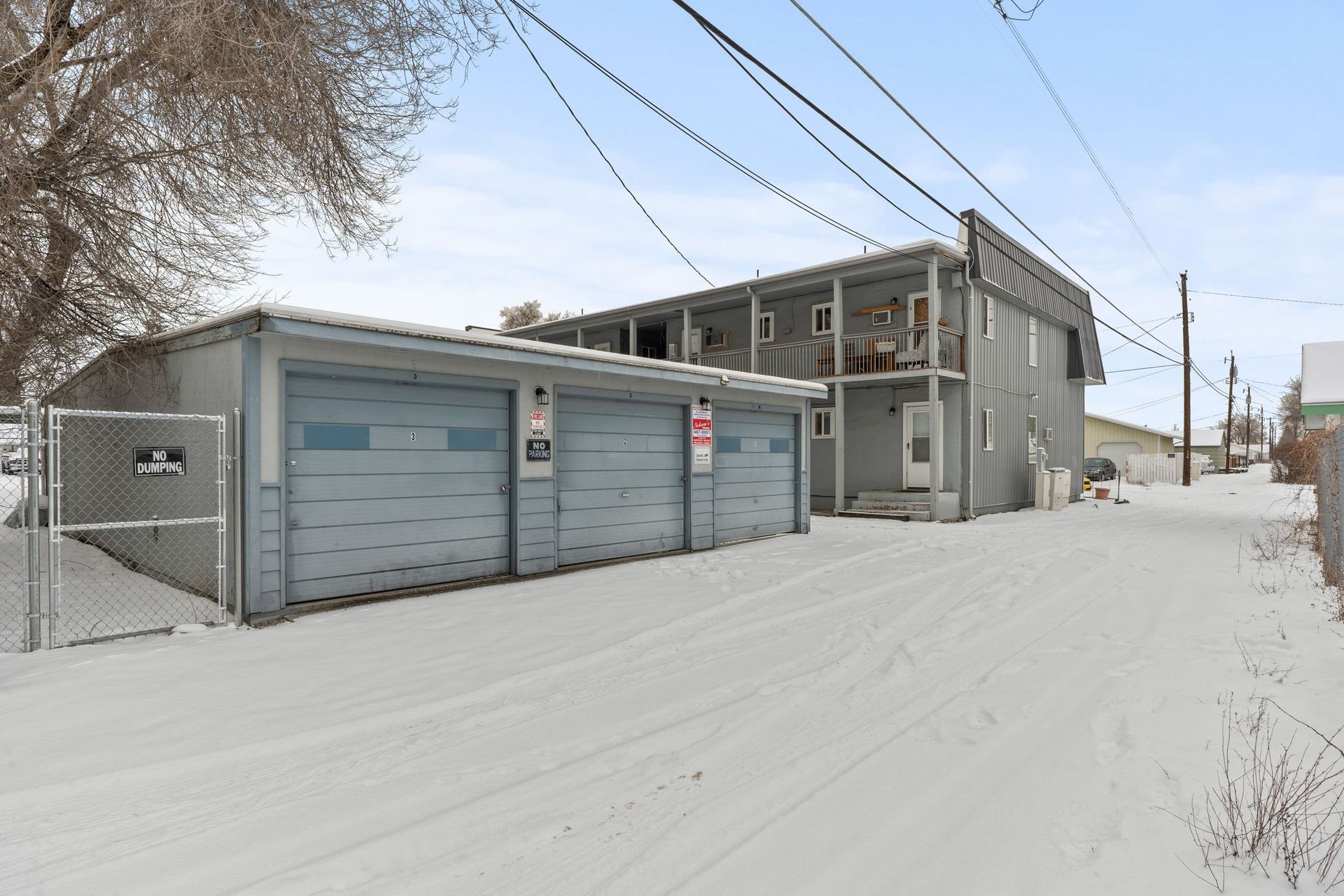 Snowy alley with a two-story gray building, detached three-car garage, and utility poles.