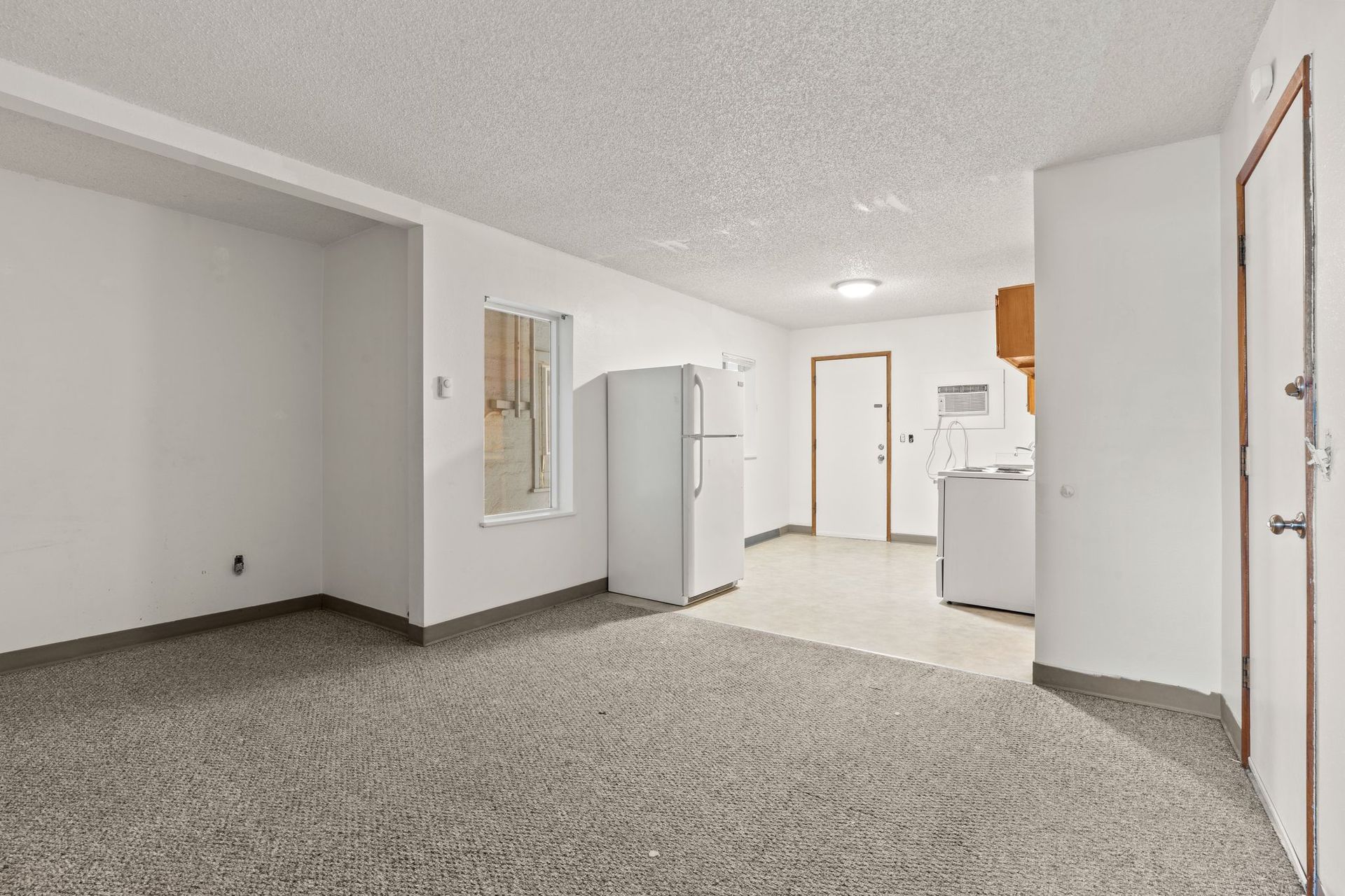 Empty room with carpet, refrigerator, and doorway to a kitchen area. White walls and ceiling.