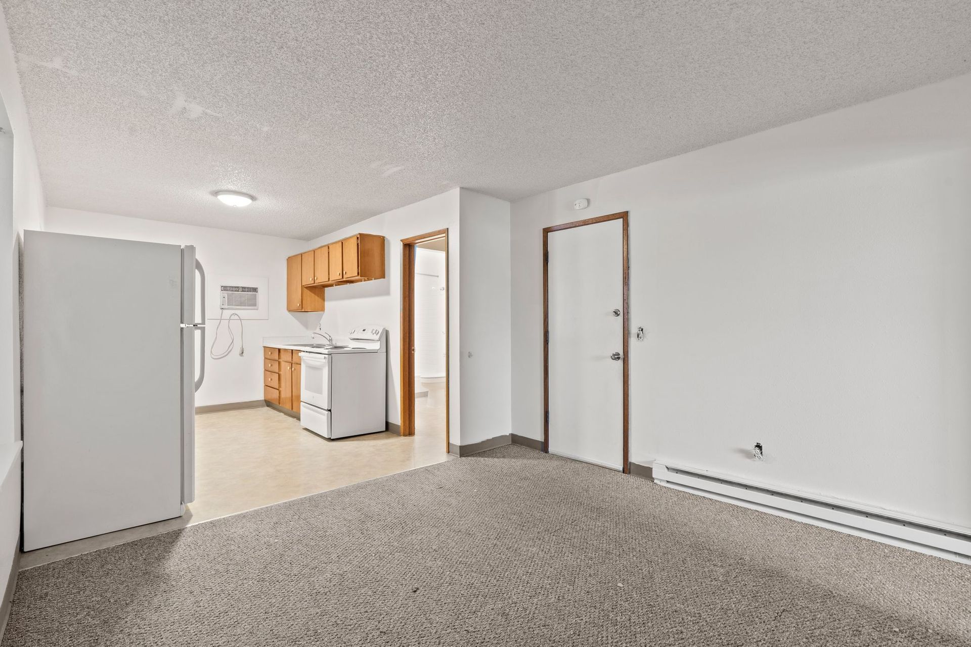 Empty apartment interior: kitchen with white appliances, wooden cabinets, and a doorway. Carpeting and white walls.