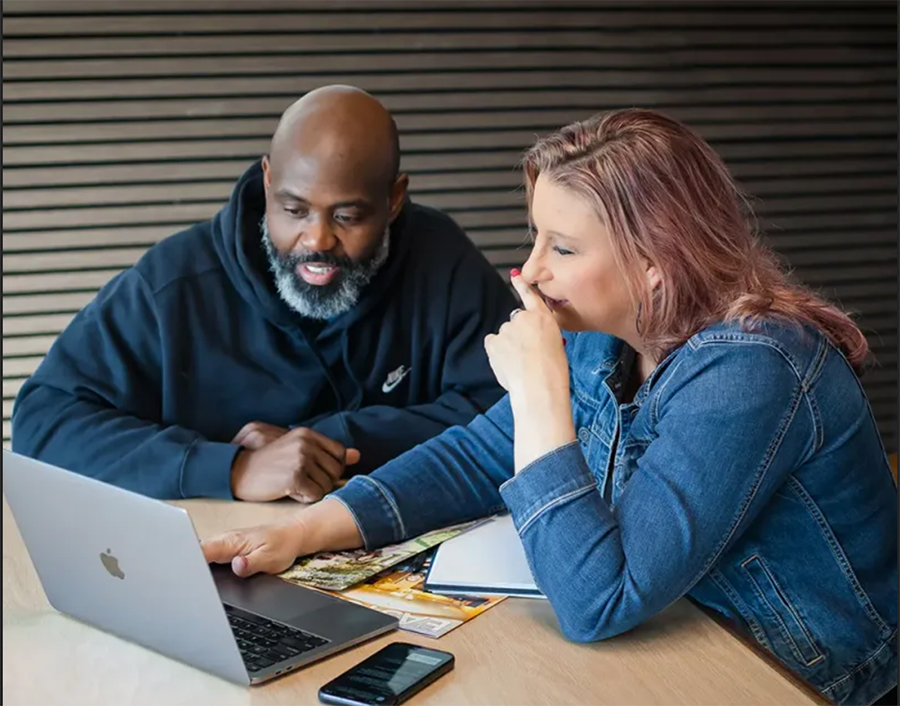 Two coworkers review a laptop at a table, discussing documents in a casual office setting.