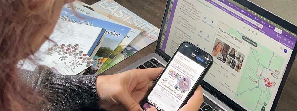 Person holding a smartphone beside a laptop displaying a website on a desk.