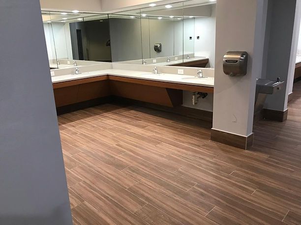 Bathroom with wood-look tile floor, large mirrors, and sinks. A paper towel dispenser and water fountain are visible.