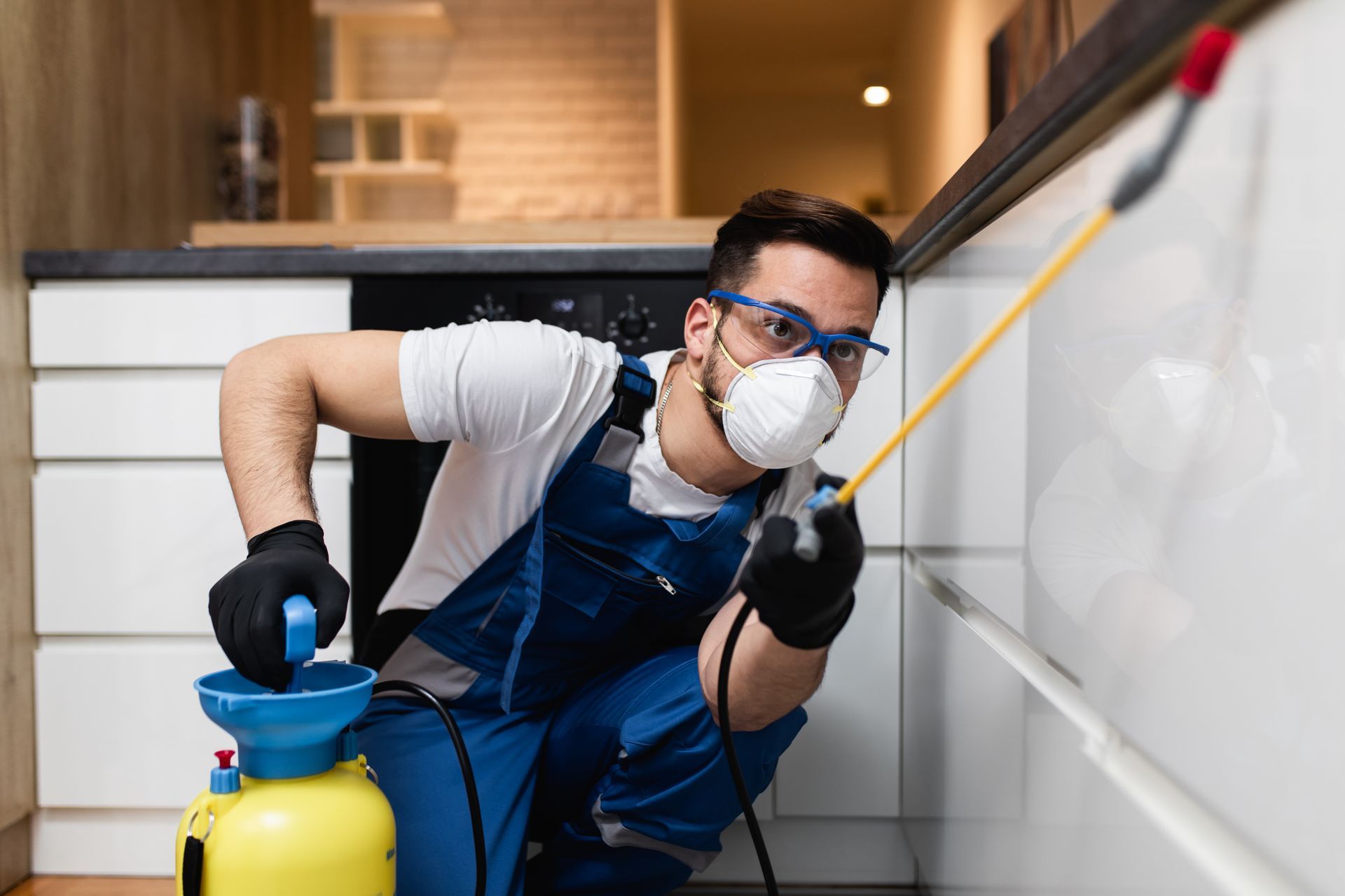 An exterminator in protective workwear is spraying pesticide in an apartment kitchen.