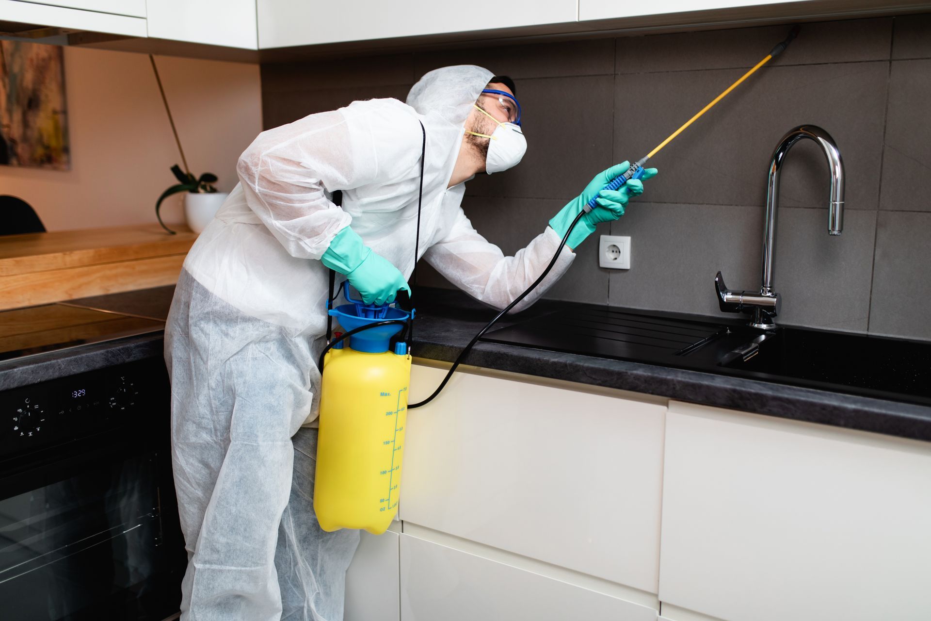 A professional exterminator in protective workwear is spraying pesticide in an apartment kitchen.