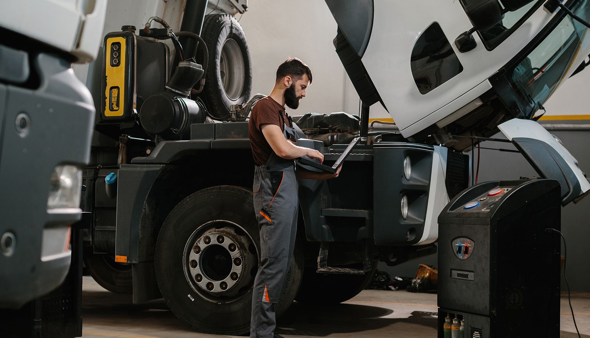 Mechanic using a laptop, checking a truck engine in a garage.