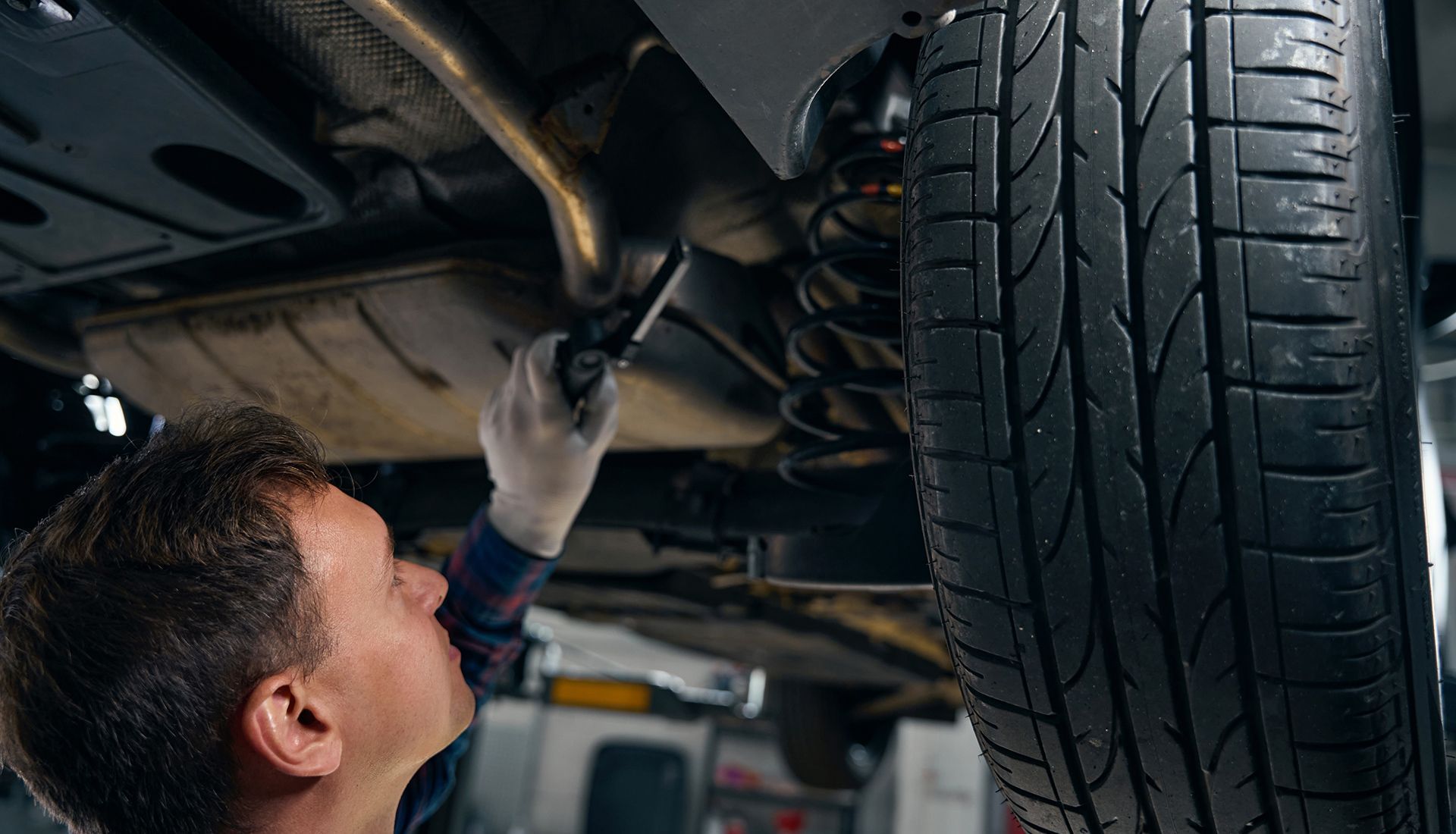 Mechanic inspecting a car's undercarriage with a wrench, tire in view; shop setting.