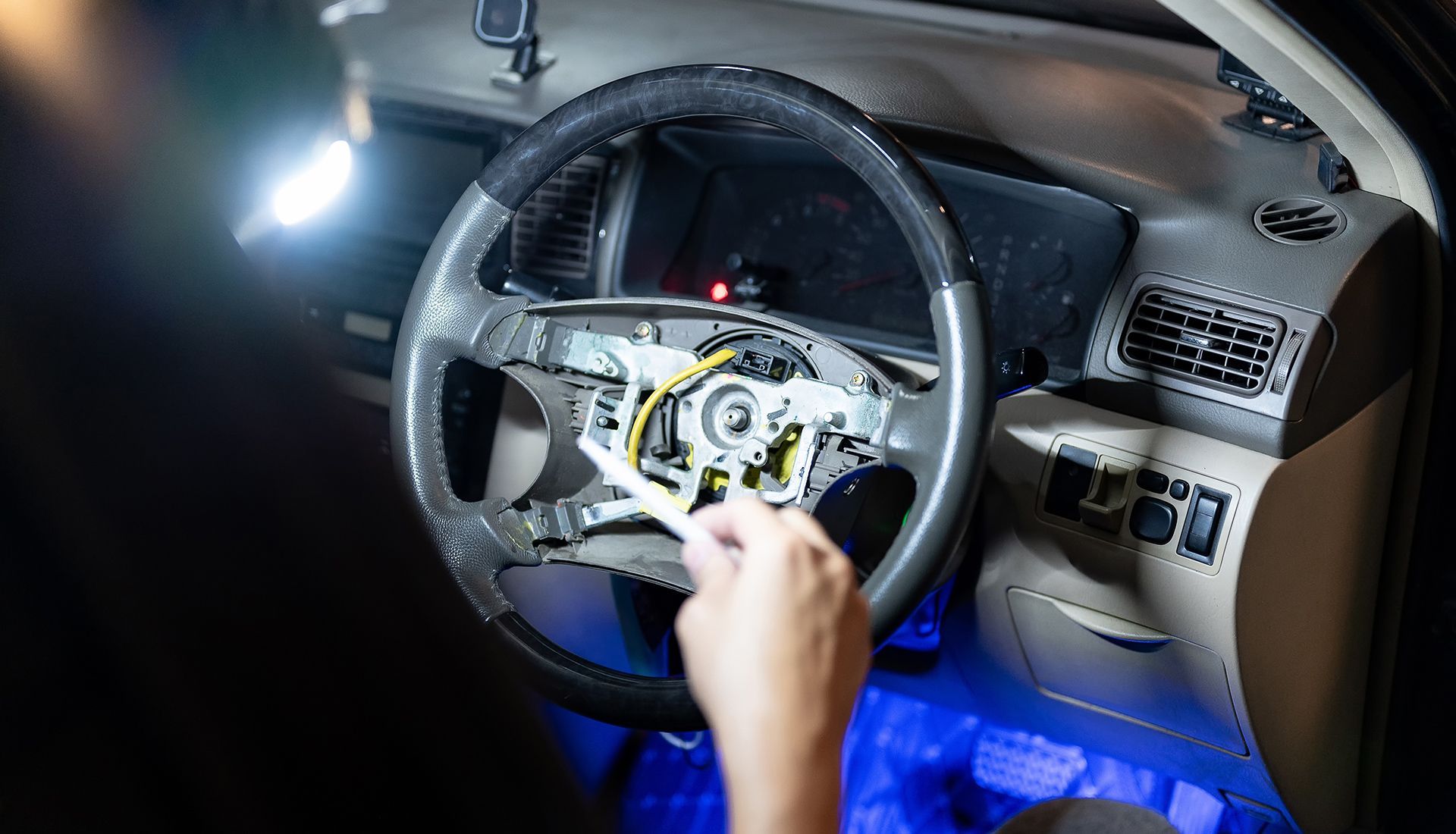 Hand working on a car steering wheel. Interior shot, wires exposed, black and gray tones.