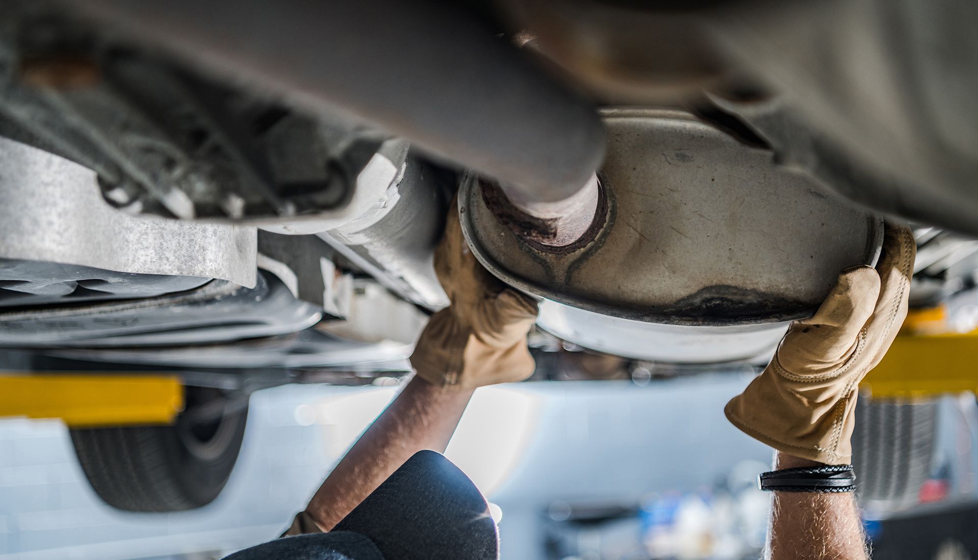Hands in work gloves, removing a car muffler from underneath a vehicle on a lift.