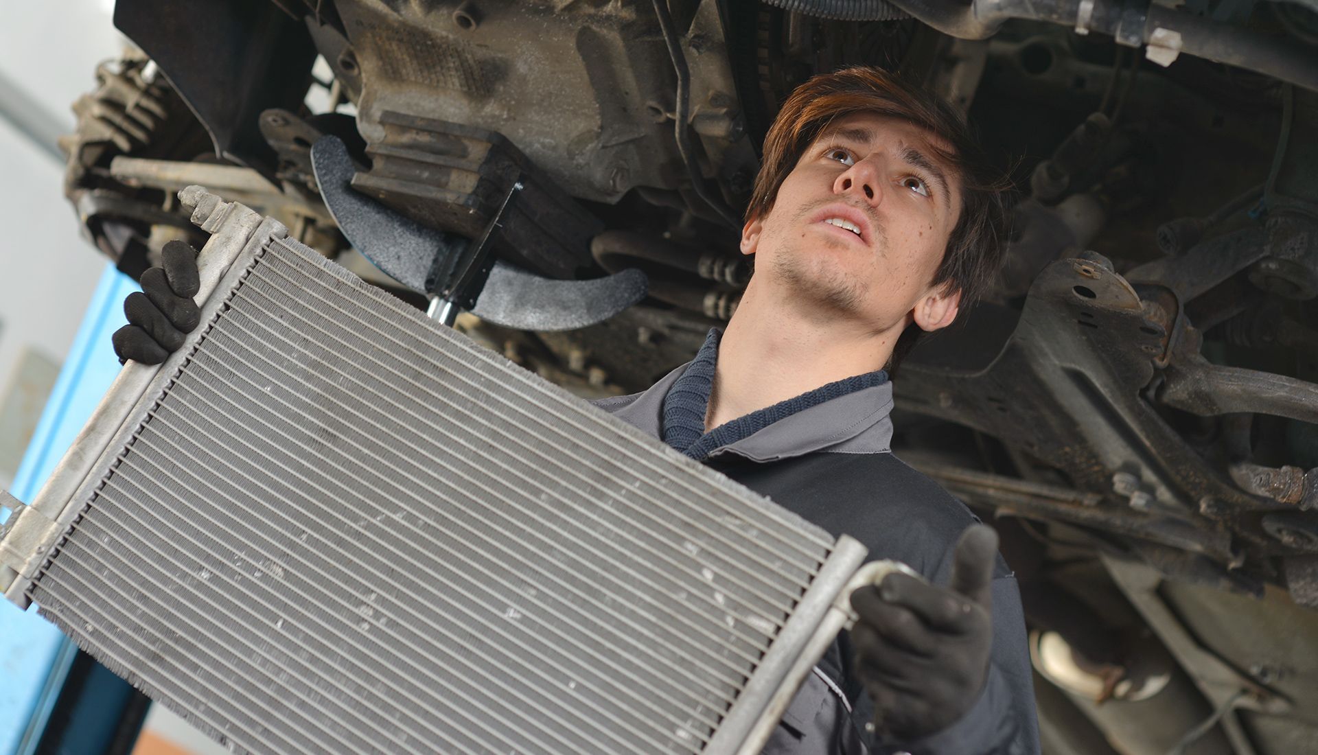 Mechanic holding a car radiator under a vehicle, looking up with a focused expression in a repair shop.