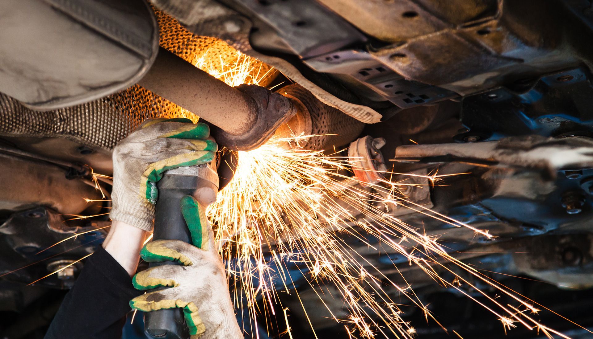 A mechanic uses a grinder to cut through a car's exhaust pipe, creating sparks.