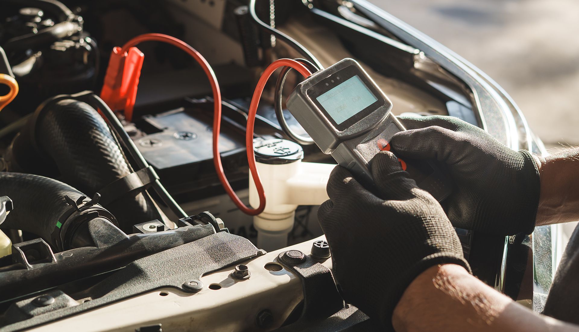 A person wearing black gloves using a battery tester connected to a car battery under the hood.