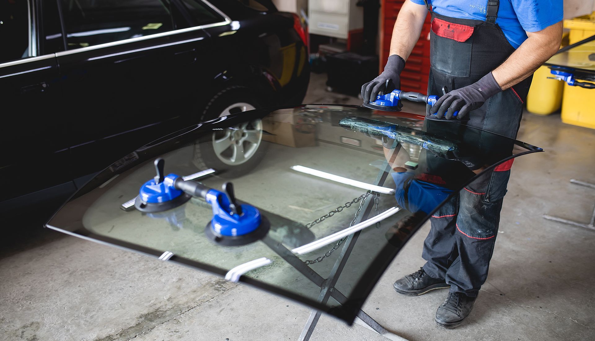 A mechanic installing a car windshield in a garage, using suction cups and tools.