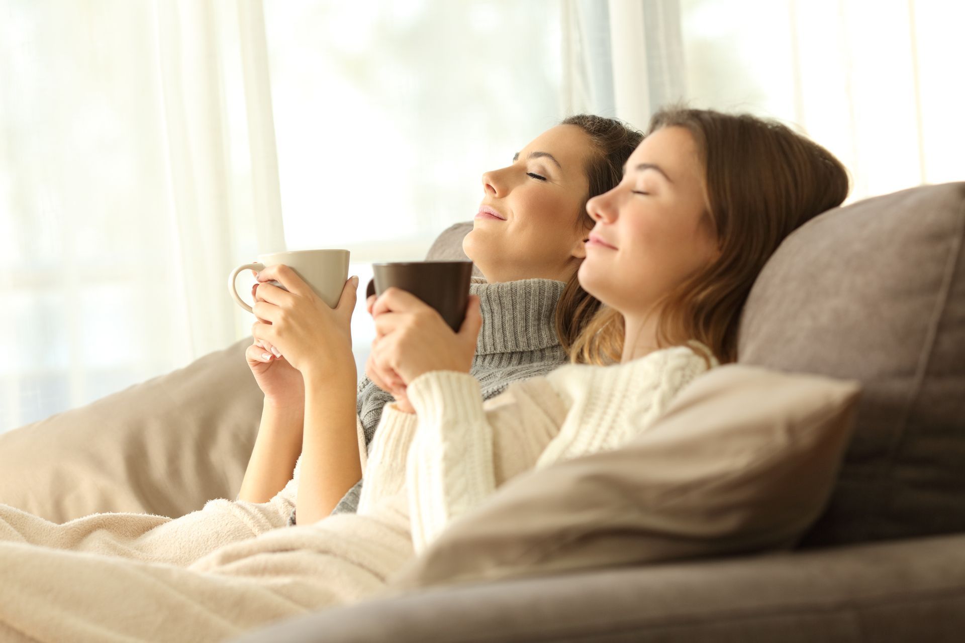 Two women are sitting on a couch holding cups of coffee.