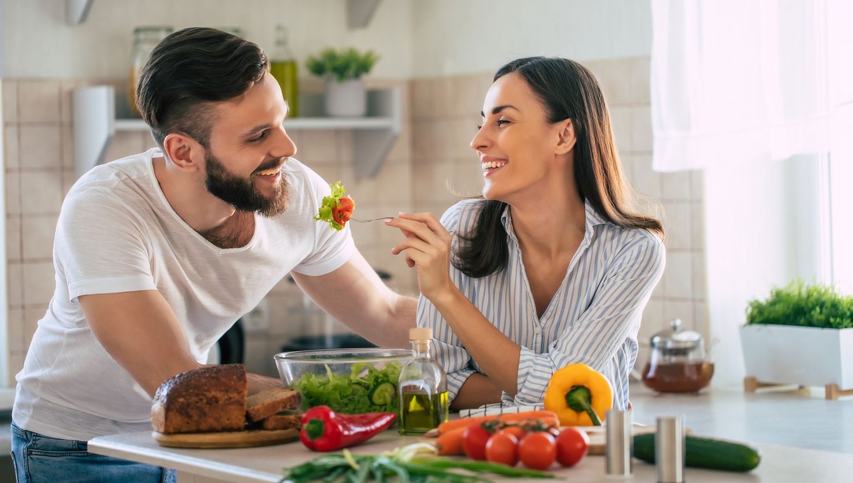 A man is feeding a woman a salad in a kitchen.