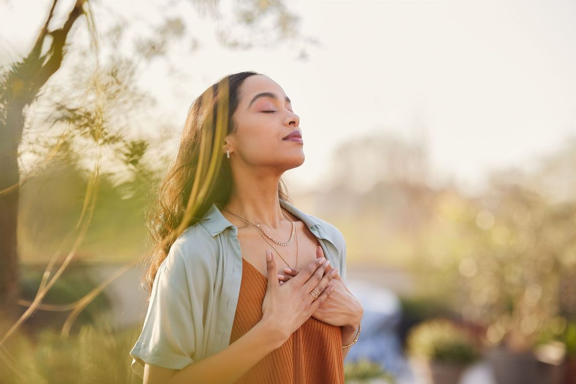 A woman is standing with her eyes closed and her hands on her chest.