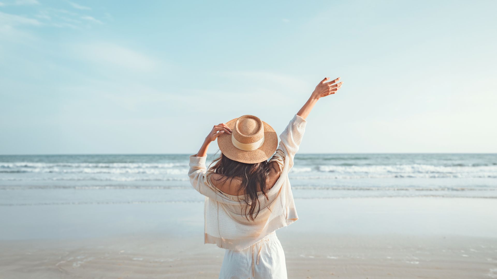 A woman wearing a straw hat is standing on the beach with her arms outstretched.