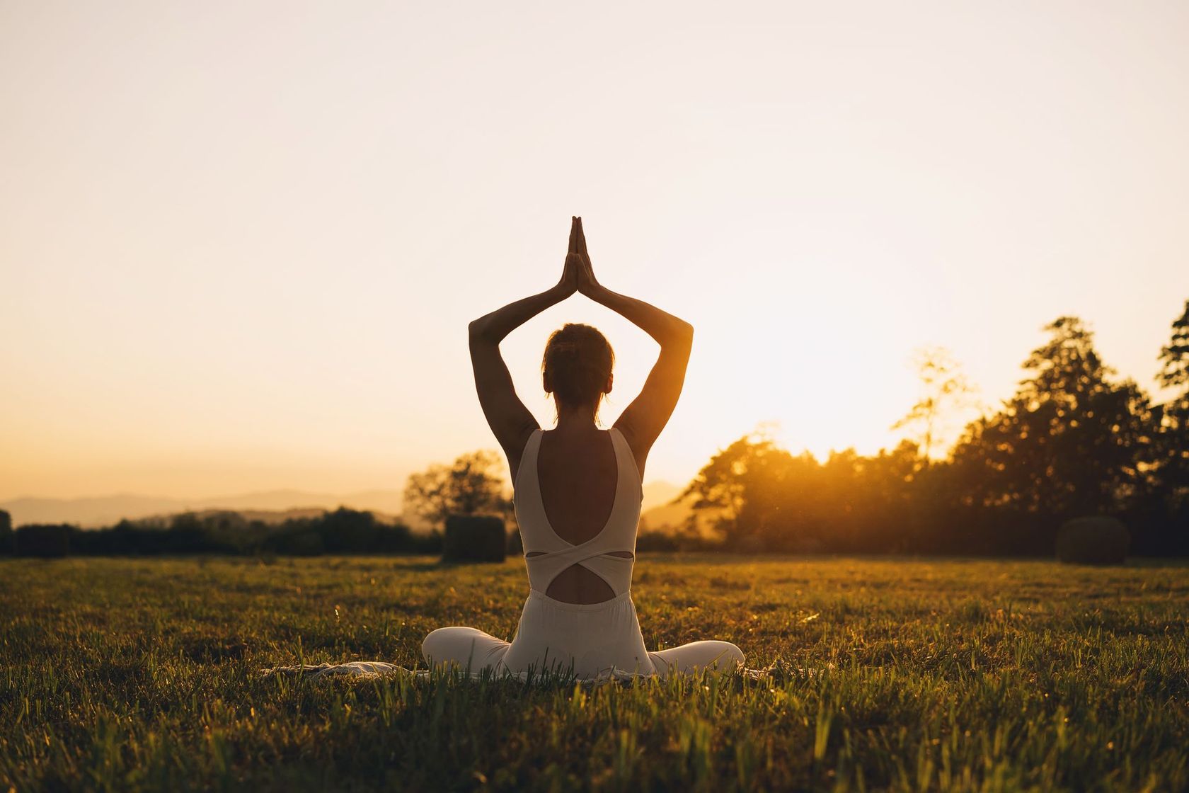 A woman is sitting in a field practicing yoga at sunset.