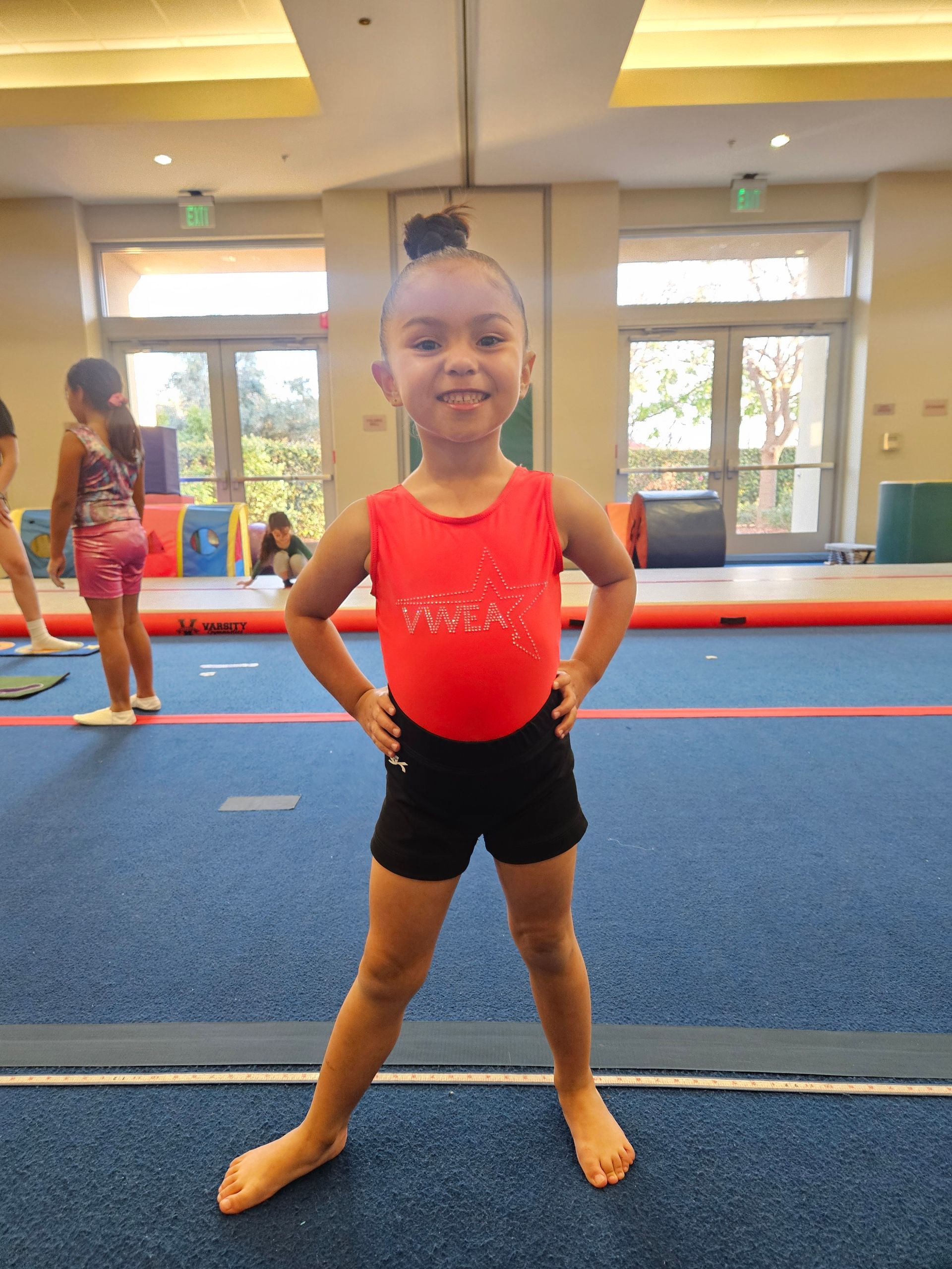Young girl in a red leotard and black shorts poses with hands on hips in a gymnastics gym.
