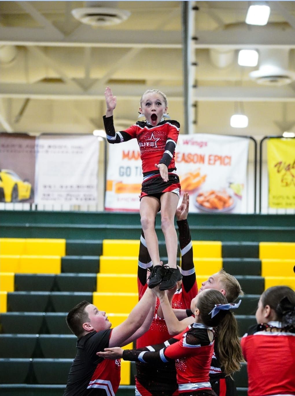 A young cheerleader is lifted into the air during a performance, with her arms raised high.