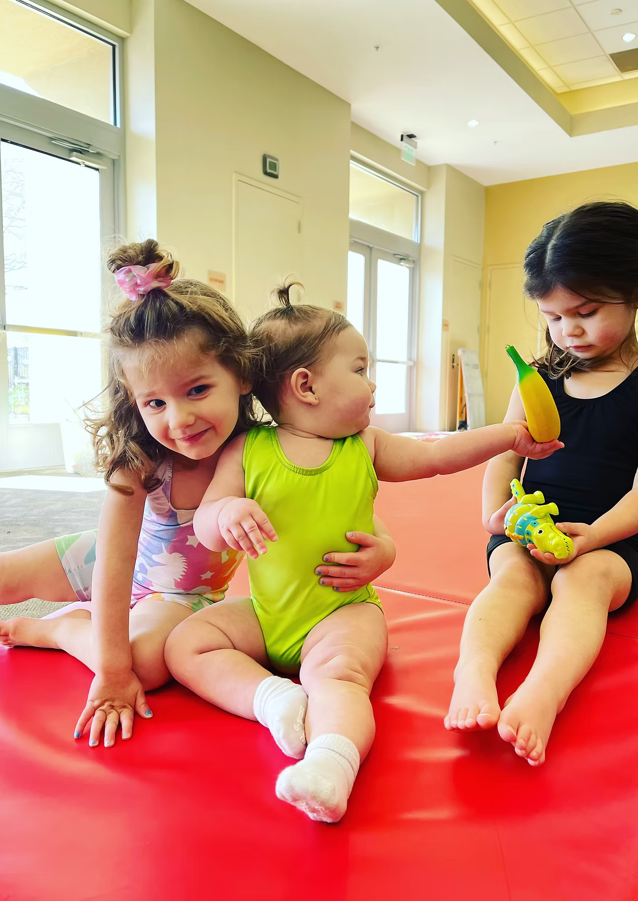 Three young children in leotards sit on a red mat, two holding toys.