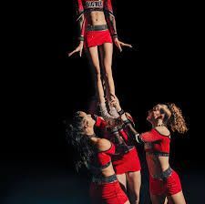 Cheerleaders performing a stunt; one person is lifted high above. Dark background; red and black uniforms.