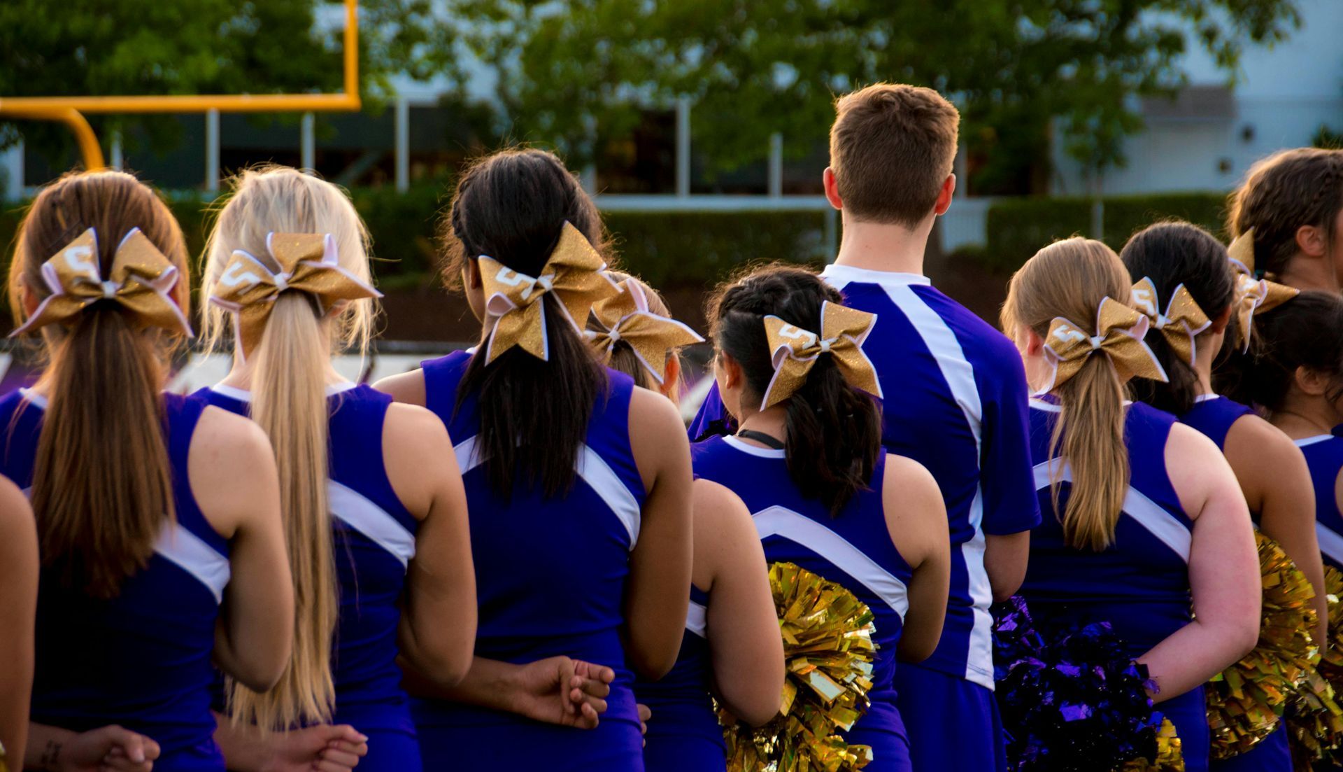 Cheerleaders in blue uniforms with gold bows stand on a field; some hold pom-poms, watching a game.