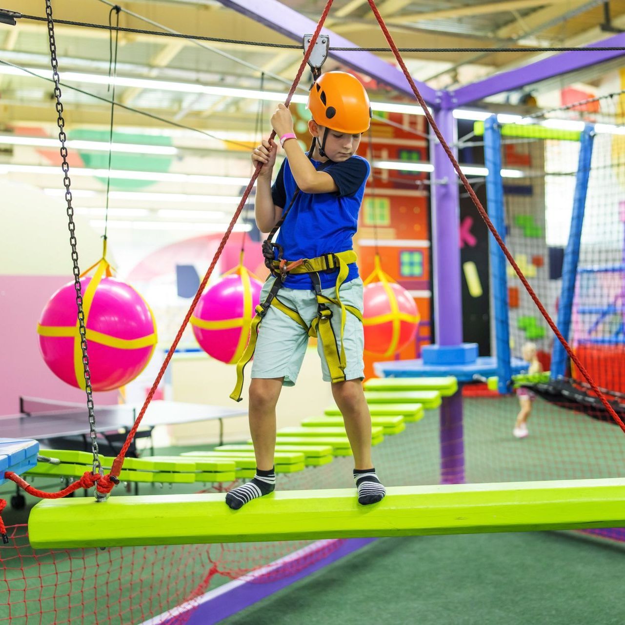 Boy in a harness navigating an indoor ropes course, balancing on a beam. He's wearing a helmet. Brightly colored setting.