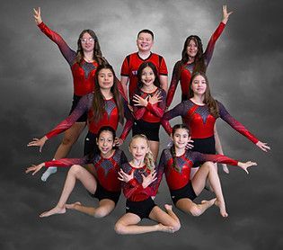 Gymnastics team in red and black leotards, posing with arms raised, smiling. Gray background.