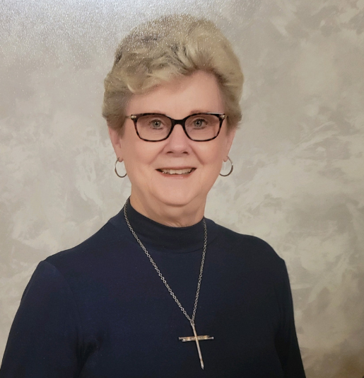 Smiling older woman with glasses, short blonde hair, wearing a blue top and cross necklace.