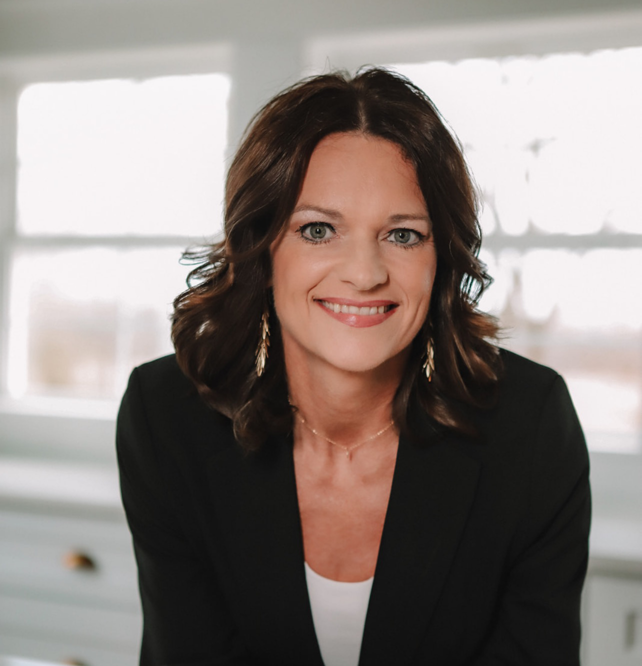 Woman with dark hair smiles, wearing a black blazer and gold jewelry, indoors in front of a window.