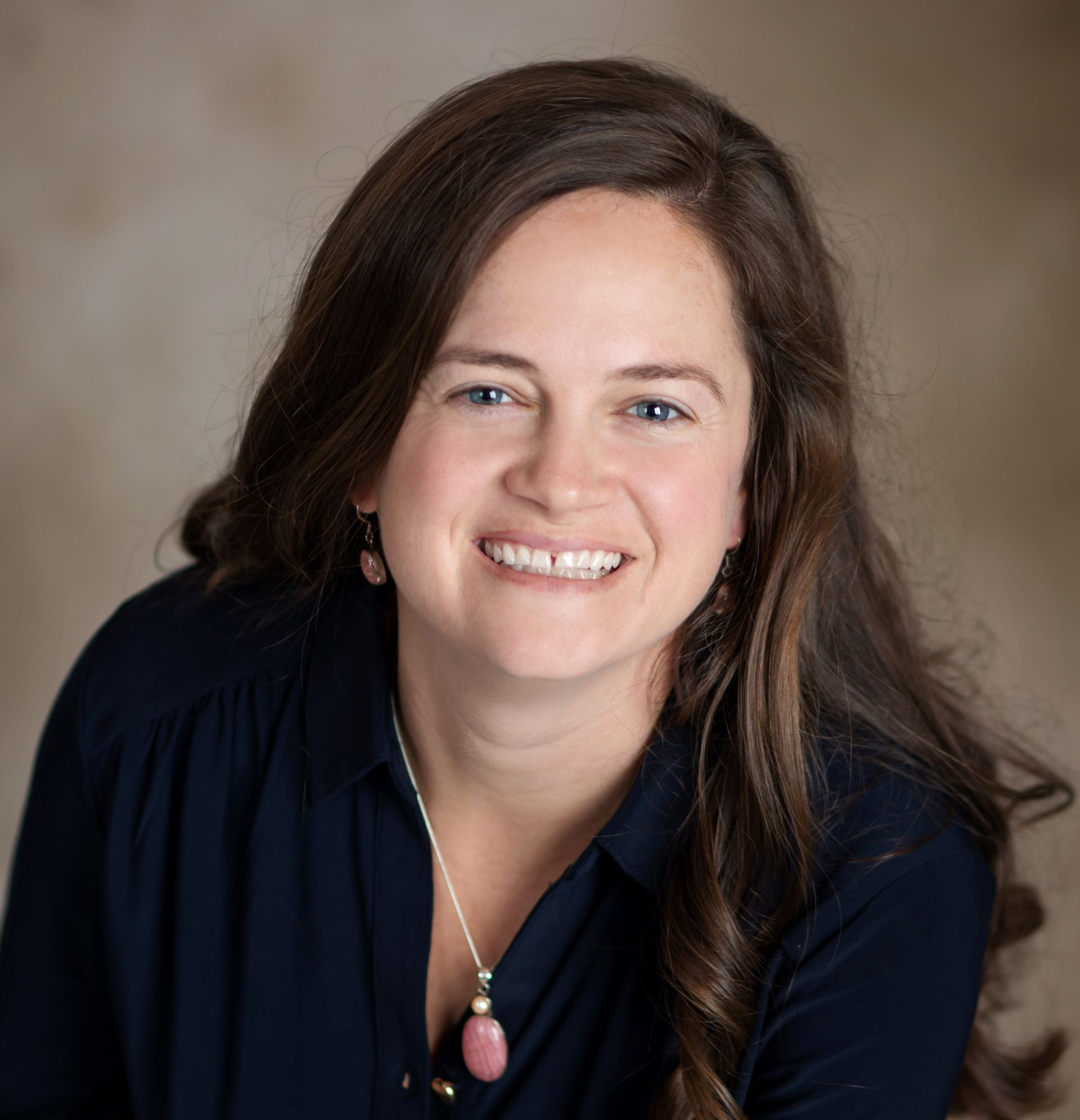 Woman with long brown hair smiles, wearing a blue blouse and necklace; light-brown background.