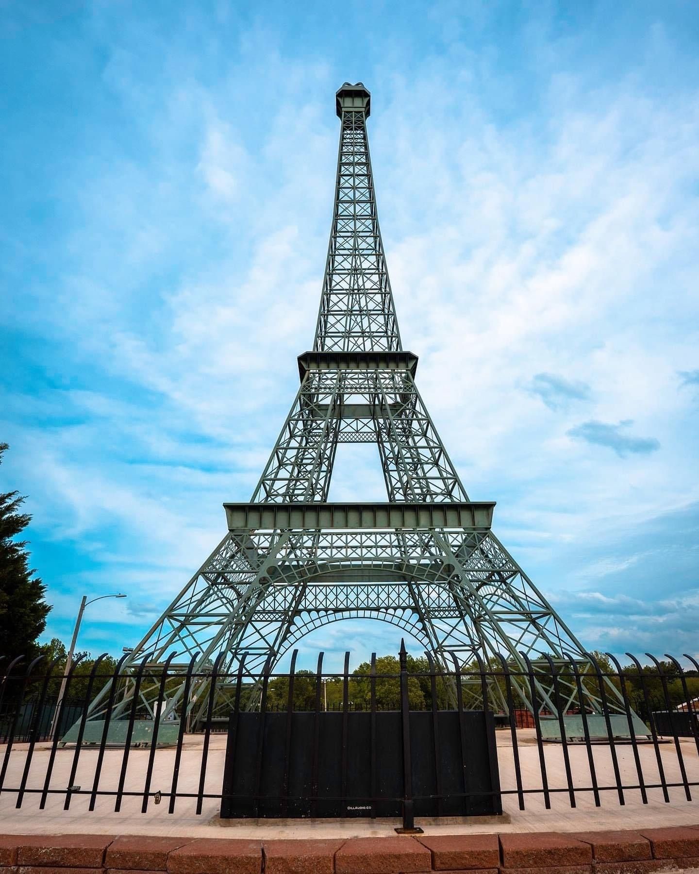 Steel Eiffel Tower replica under a blue sky with clouds, enclosed by a black fence.