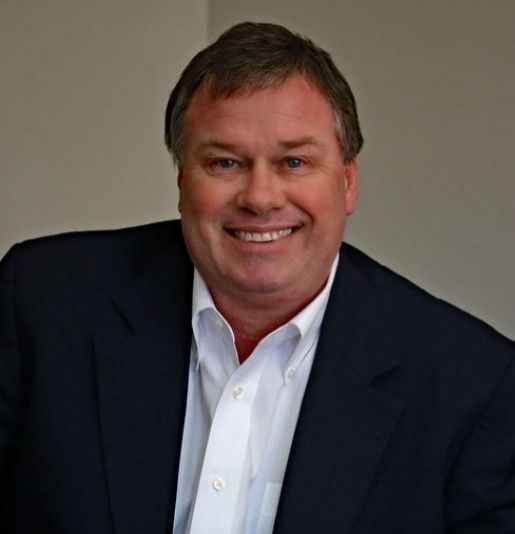 Middle-aged man in a dark blazer and white shirt smiles, indoors with a neutral background.