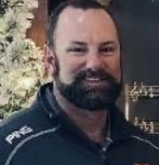 Man with a full beard, smiling. Wearing a dark shirt with a