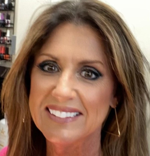 Woman with brown hair, dark eye makeup, smiling, wearing gold earrings; indoors.