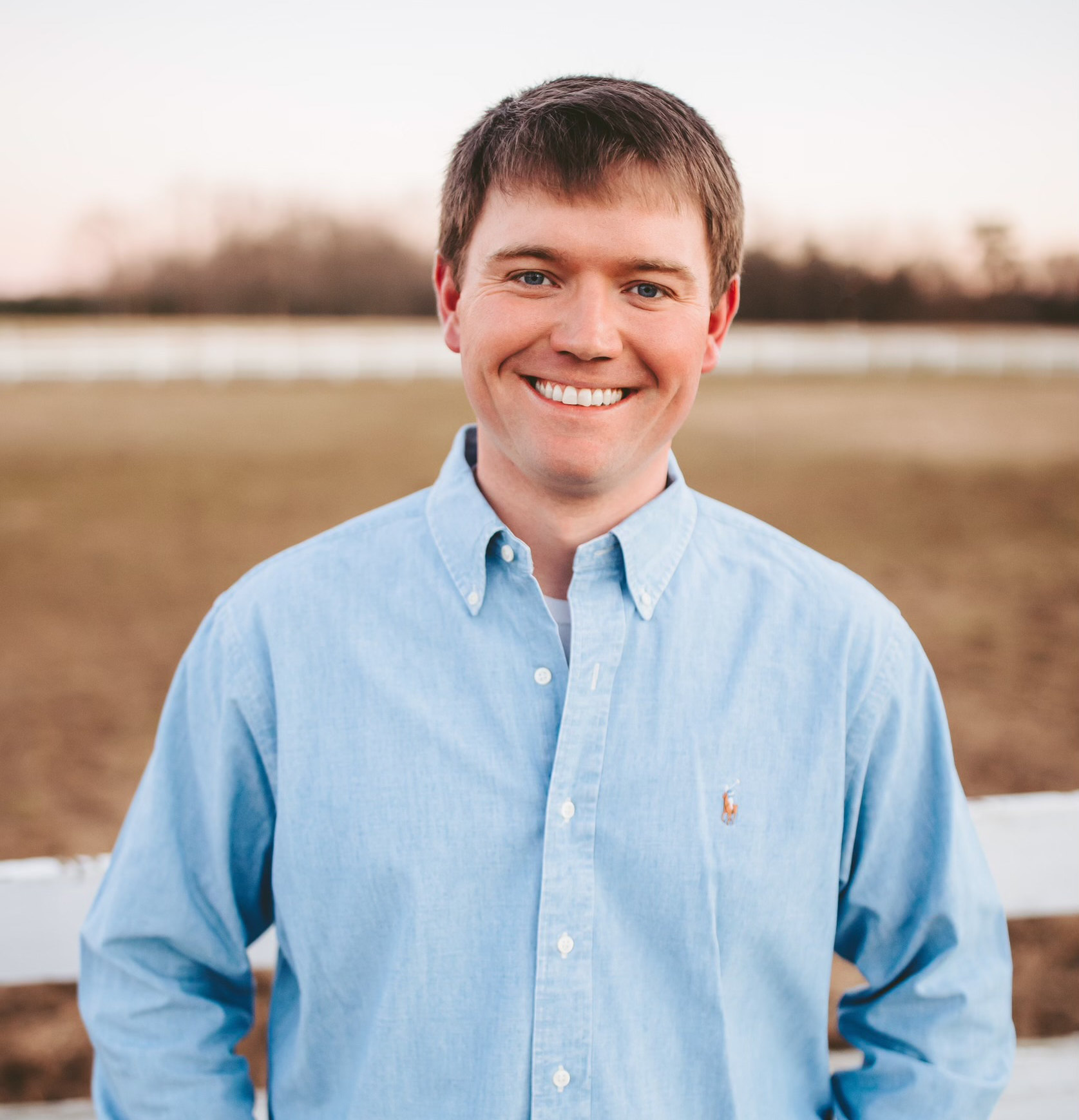 Man smiling, hands in pockets, wearing a blue button-down shirt, standing outside near a fence and field.