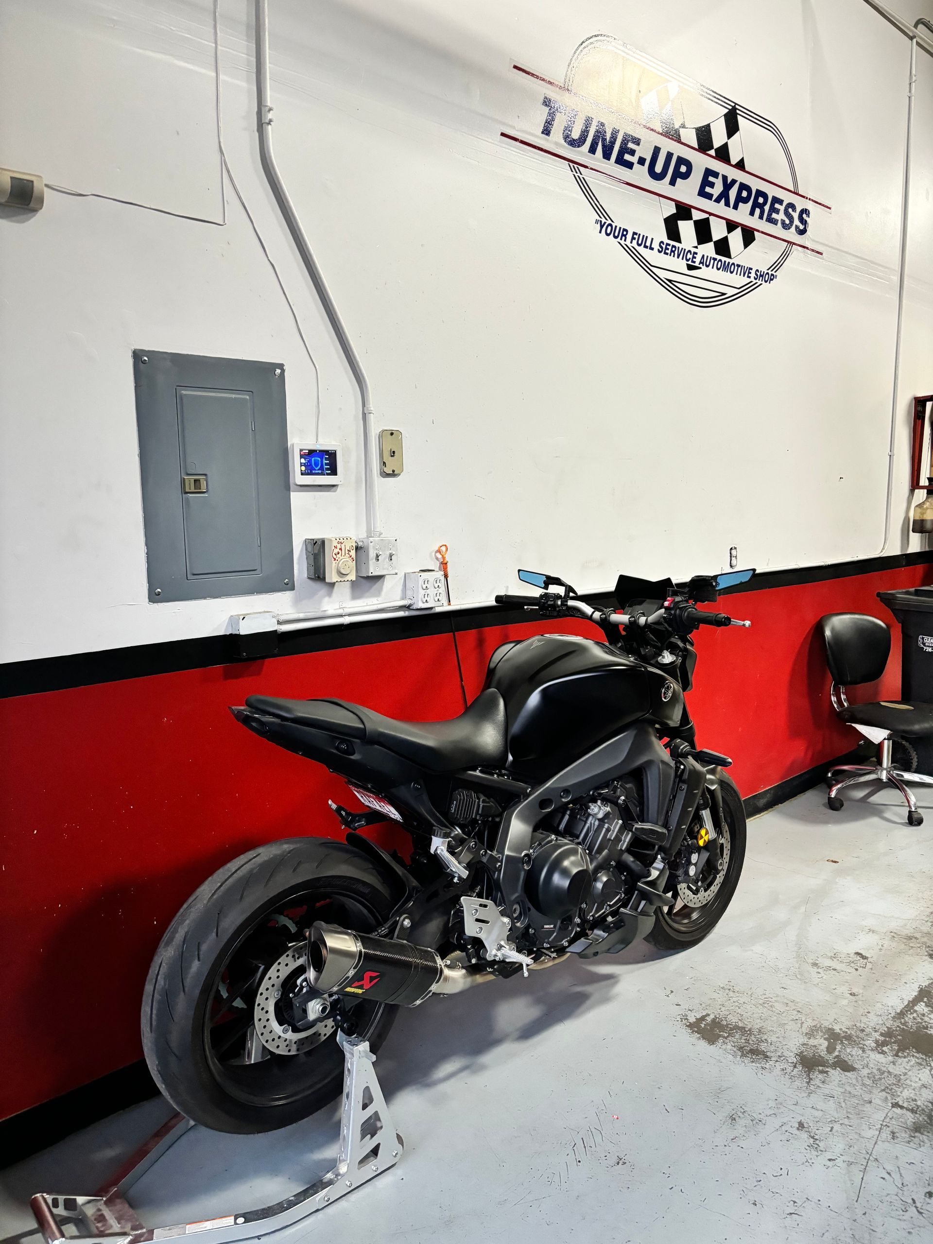 Black motorcycle parked indoors against a white wall with red trim and a logo sign above. | Tune-Up Express Auto