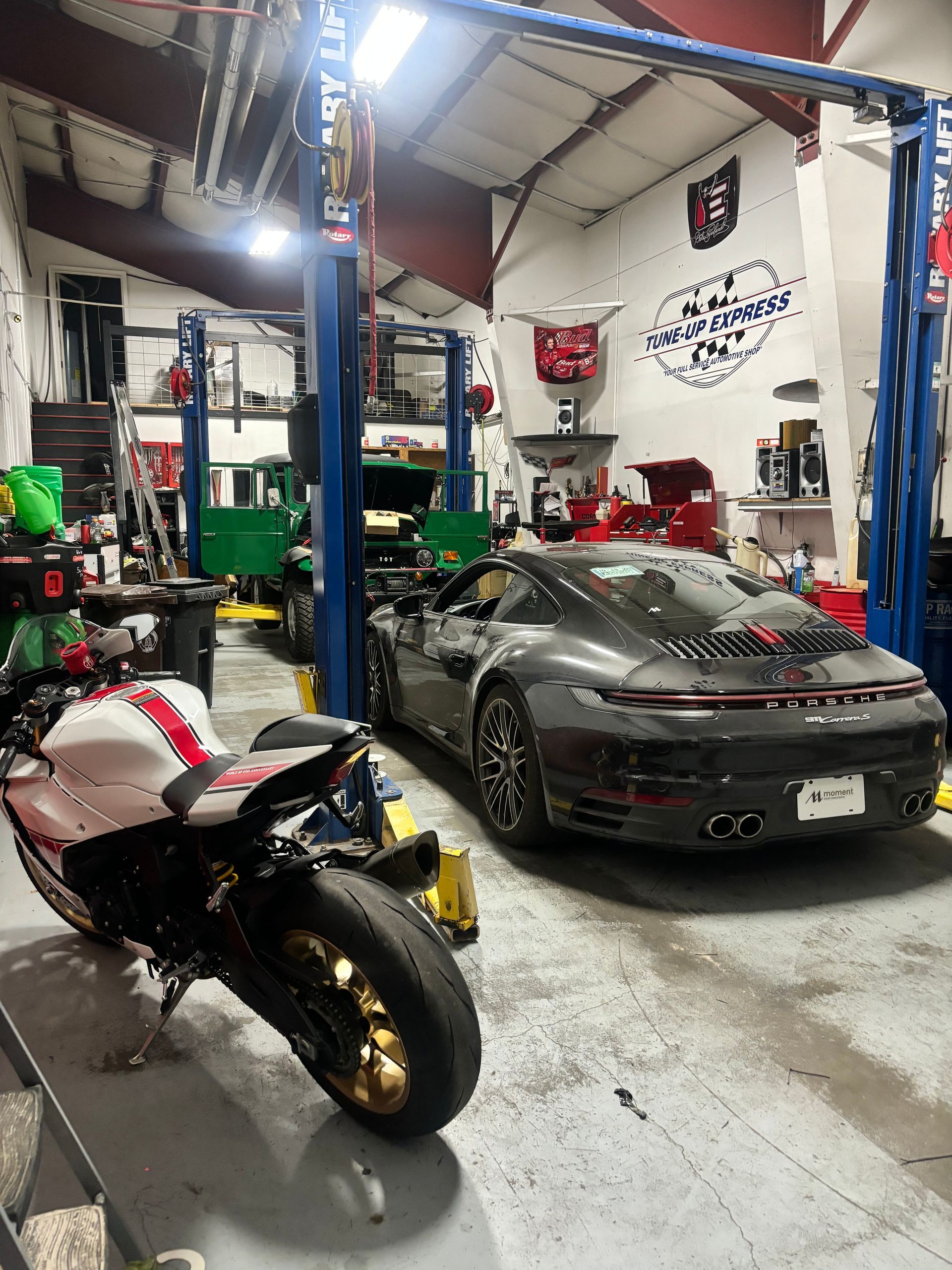 Motorcycles and a sports car inside a cluttered auto repair garage with a lift | Tune-Up Express Auto