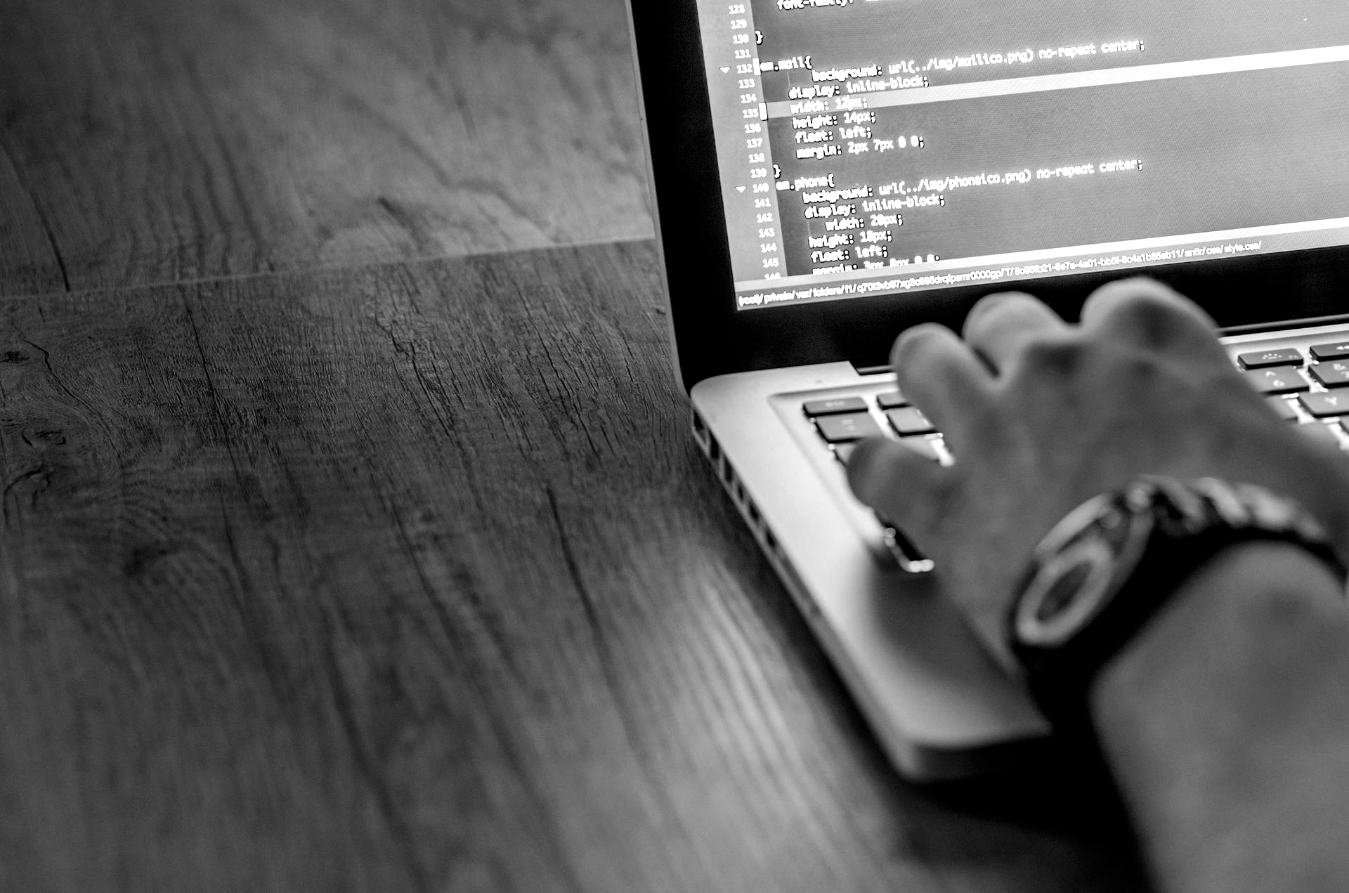 Una persona escribiendo código en una computadora portátil; escritorio de madera, reloj en la muñeca, blanco y negro.