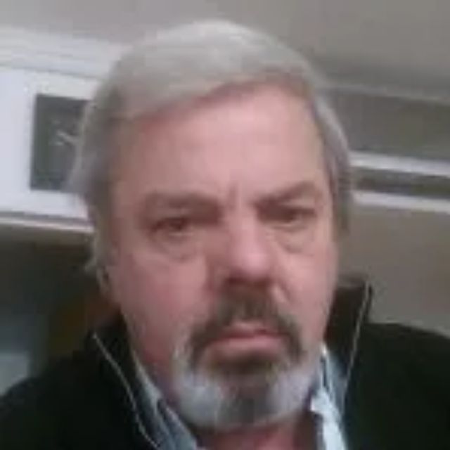 Hombre de cabello y barba grises, mirando directamente a la cámara, vistiendo una chaqueta negra sobre una camisa azul claro.