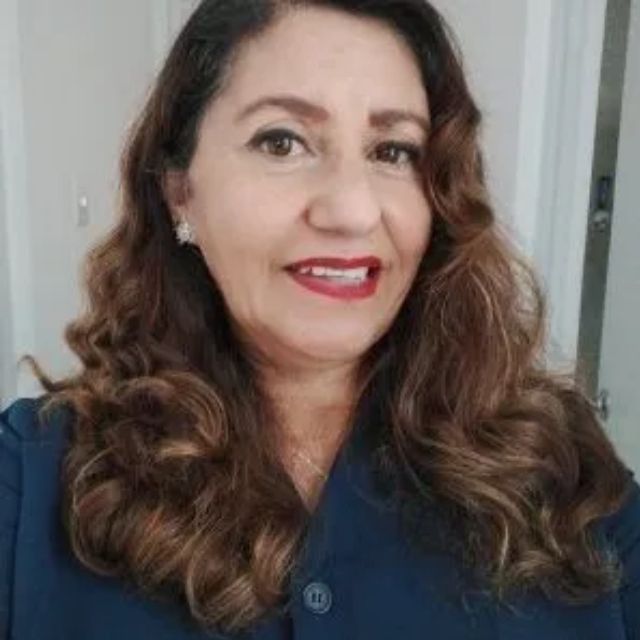 Mujer con cabello castaño ondulado, vistiendo un top azul oscuro, sonriendo con lápiz labial rojo.