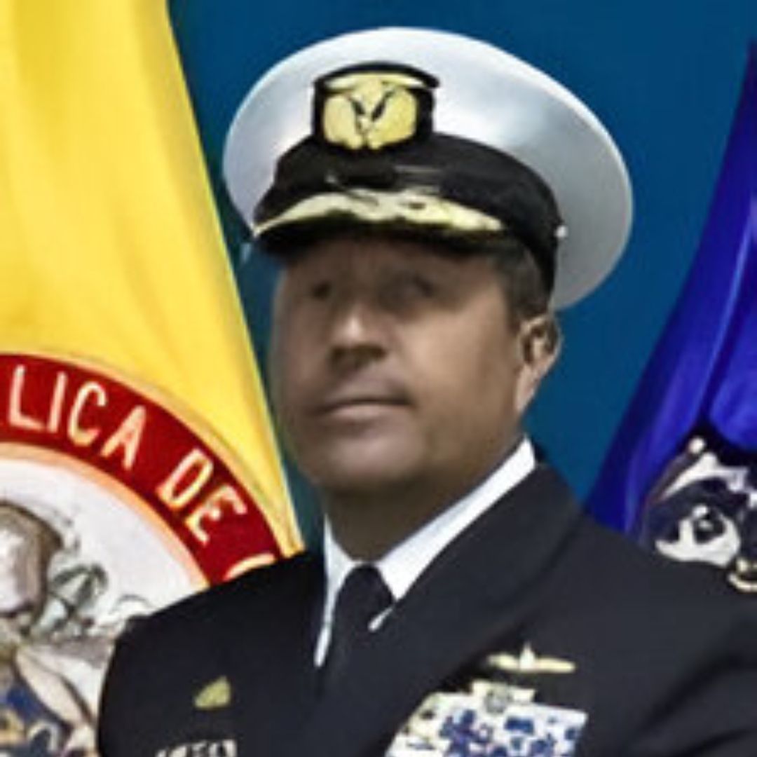 Un oficial naval uniformado con medallas, de pie frente a banderas.