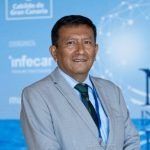 Hombre con traje claro y corbata, sonriendo, delante de un fondo azul y blanco con texto.