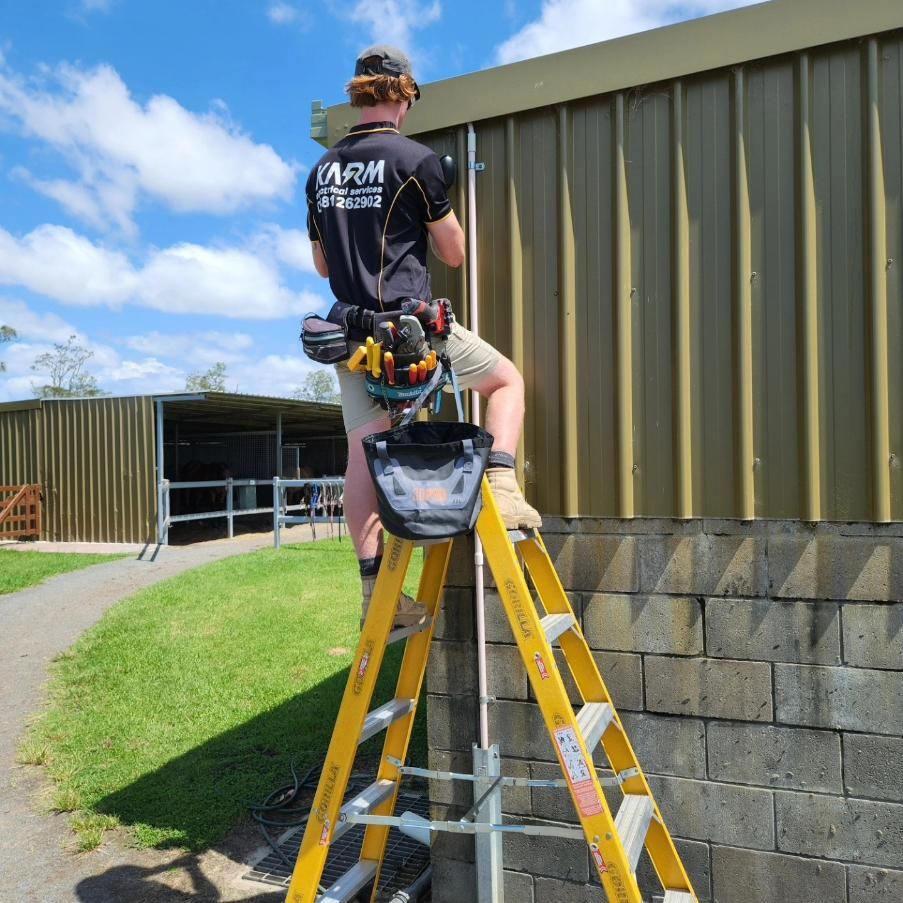A Man Standing on A Yellow Ladder with The Word Arm on His Back — KARM Electrical Services In Seaham, NSW