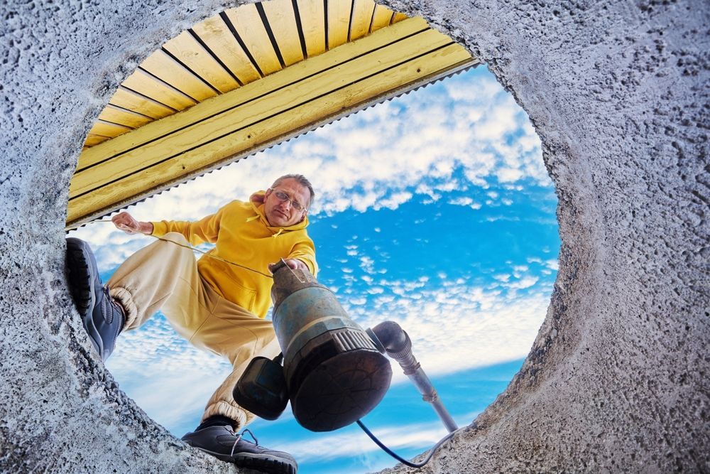 Man looking down through a circular opening, holding a tool. Yellow hoodie, blue sky.