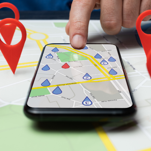 A hand touches a phone screen displaying a map with location pins; red and blue pins on the map and around the phone.