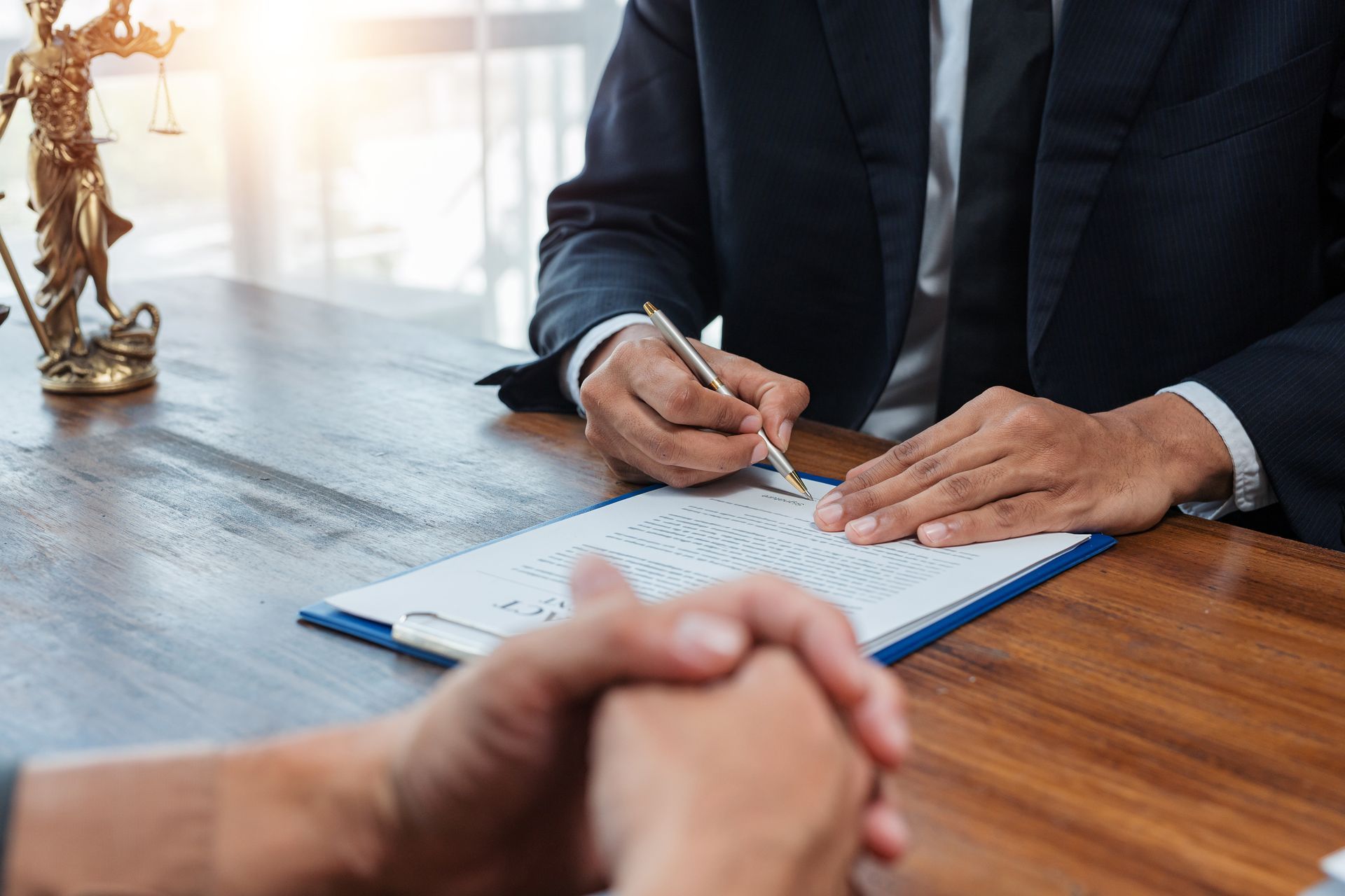 A person signing a document on a table, with another person's hands clasped in front. Legal setting A person signing a document on a table, with another person's hands clasped in front. Legal setting